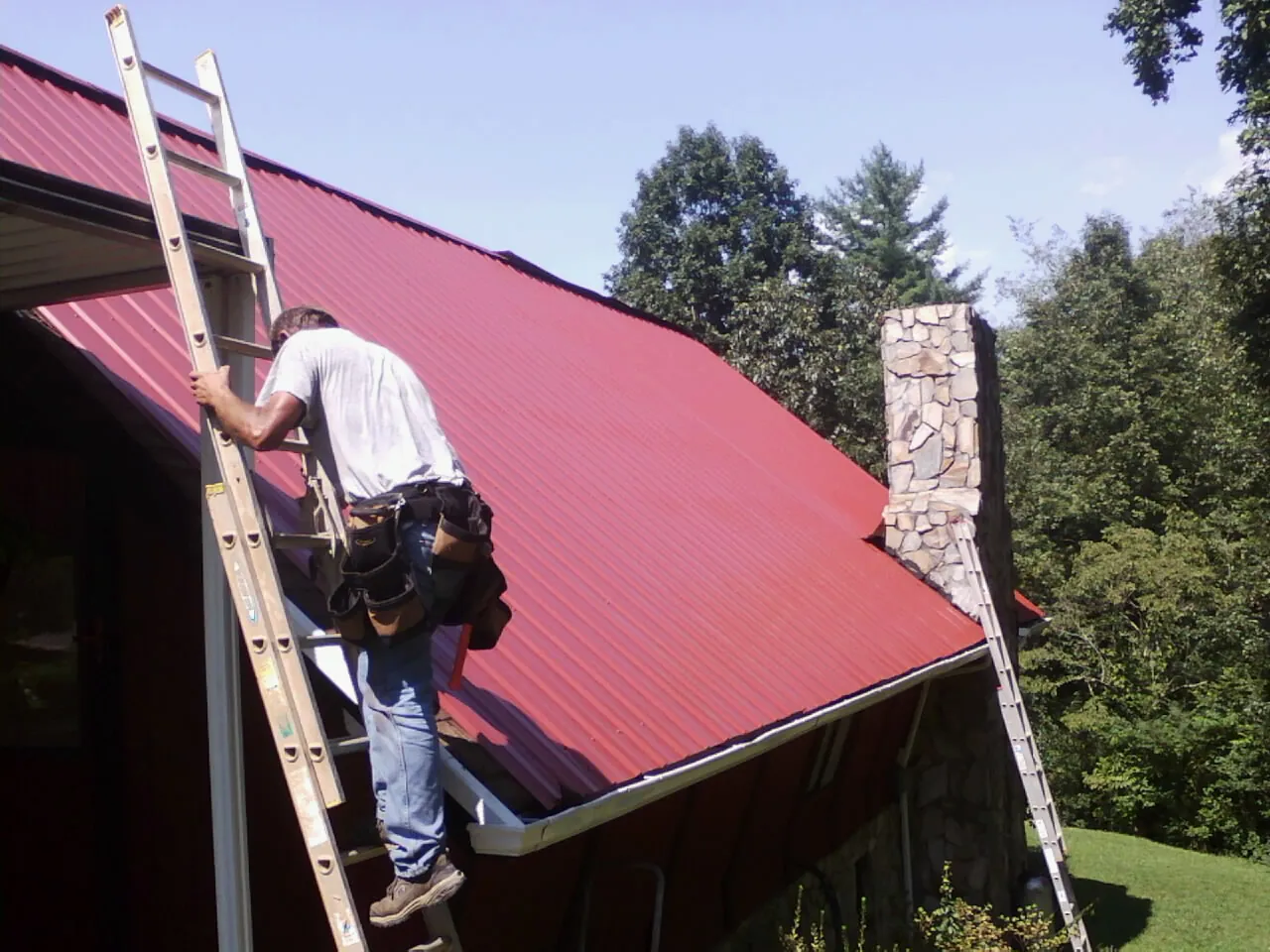 Roofer on a ladder beside a red metal roof and stone chimney — Soffit & Fascia Repair by Roofing Brothers in Cassandra